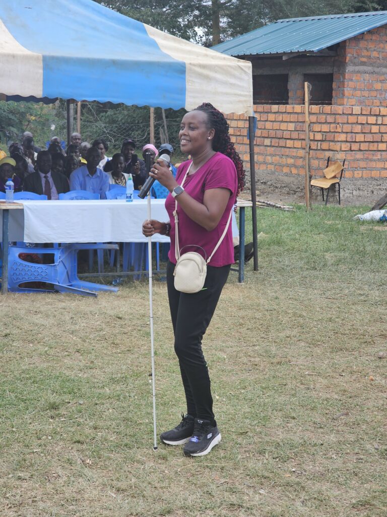 Loice Bunei speaking to the attendees at Korara’s celebration of White Can Day. She stands in front of other members of the CPI team in the mid-afternoon sun.